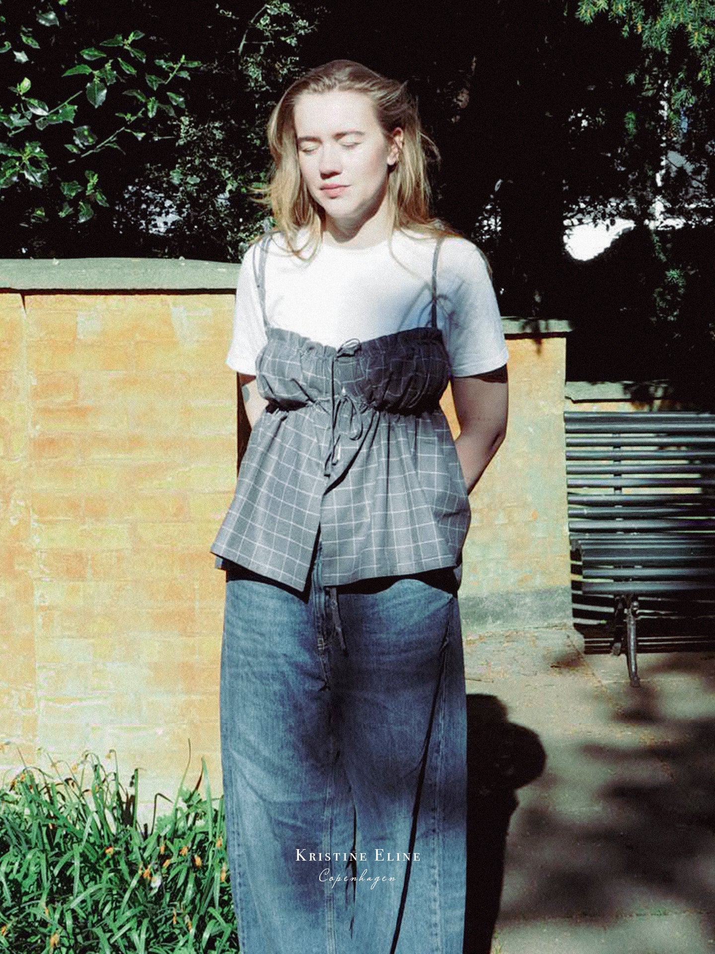 Model wearing a grey checkered ruffle top and blue jeans standing outdoors with a bench and wall in the background.