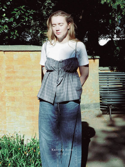 Model wearing a grey checkered ruffle top and blue jeans standing outdoors with a bench and wall in the background.
