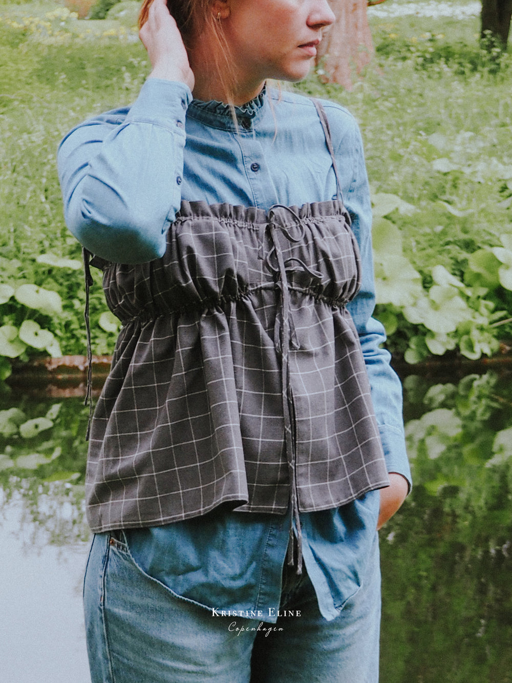 Person wearing a grey checkered ruffle top over a blue shirt with a blurred green natural background