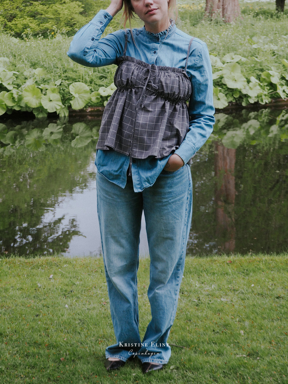 Person wearing a blue denim outfit with a grey checkered ruffle top on top by a pond