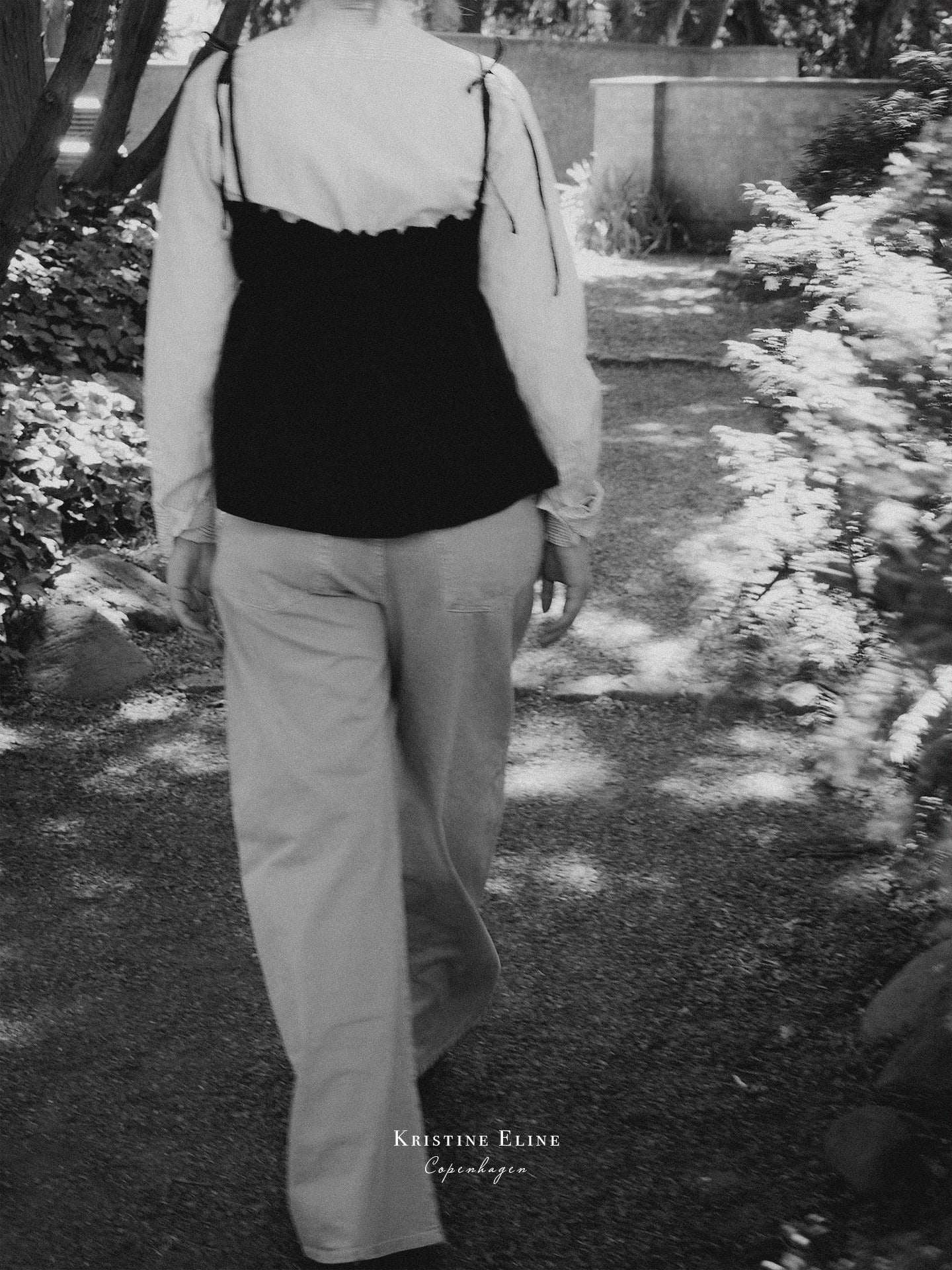 A black and white photograph of a person from behind, wearing a black top over a white long-sleeve top and light-colored trousers, walking along a garden path. The text 'Kristine Eline' and 'Copenhagen' is visible at the bottom.