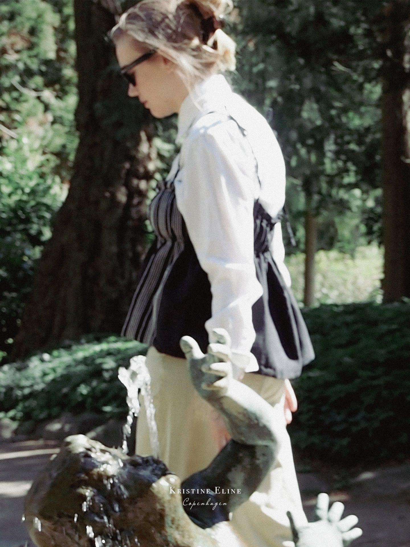 A woman in a white shirt with a black top on top and beige pants. She stands outdoors near a fountain in a park, wearing dark sunglasses. A small watermark at the bottom reads 'KRISTINE ELINE COPENHAGEN'.