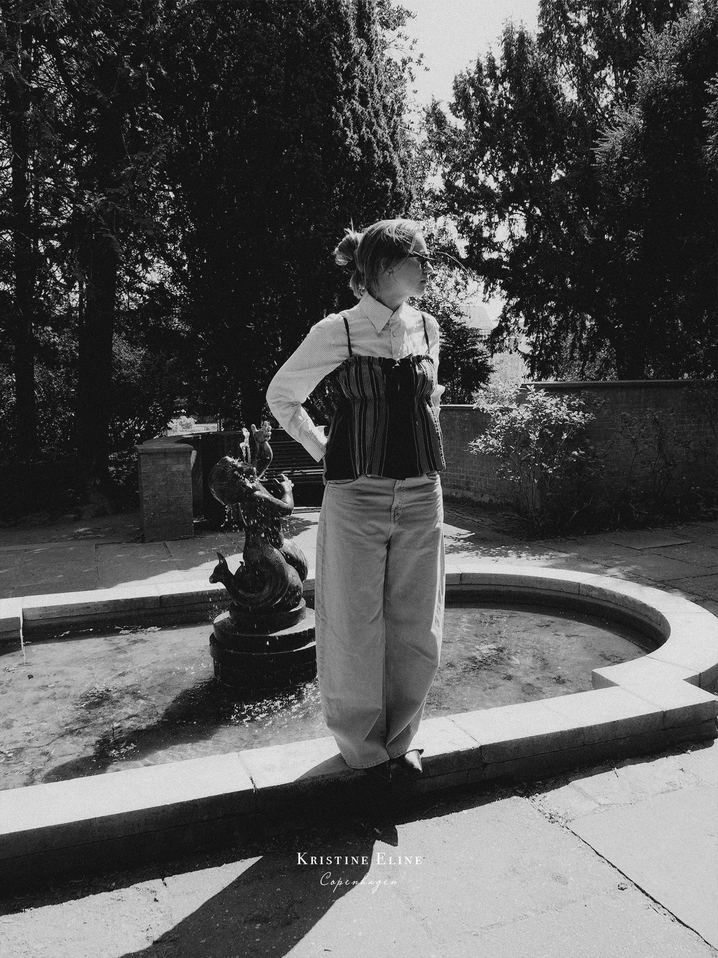 A black and white fashion photograph of a model standing outdoors by a stone fountain, wearing a white shirt, a striped black top with thin straps, and light-colored wide-leg trousers. Trees and a stone wall are visible in the background.