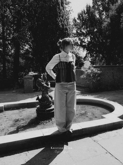 A black and white fashion photograph of a model standing outdoors by a stone fountain, wearing a white shirt, a striped black top with thin straps, and light-colored wide-leg trousers. Trees and a stone wall are visible in the background.