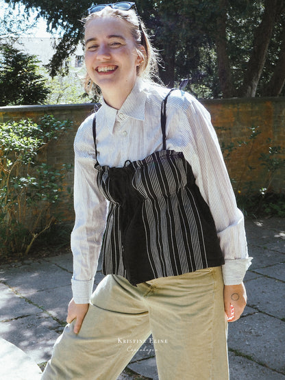 A woman outdoors wearing a white and black vertically striped long-sleeve button-up shirt layered under a black ruffled top with thin black straps, paired with beige pants.