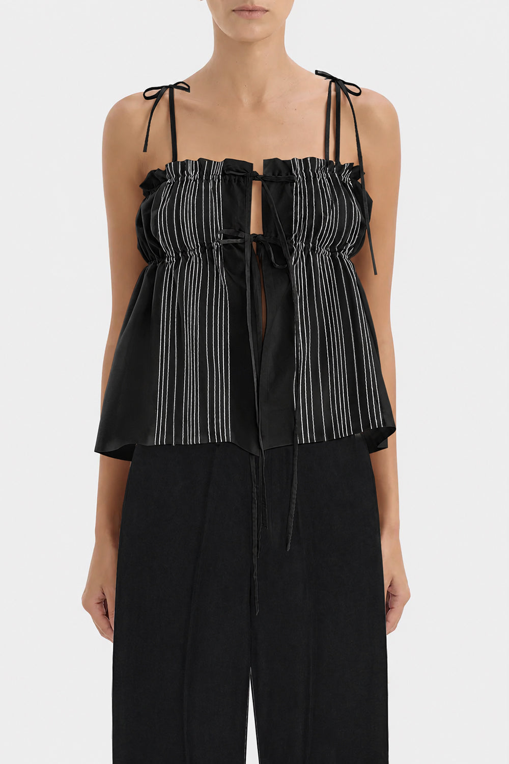 A model wearing a black top with white vertically stripes on the front with thin shoulder ties and black pants, shown against a light gray studio background.