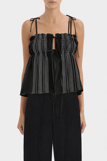 A model wearing a black top with white vertically stripes on the front with thin shoulder ties and black pants, shown against a light gray studio background.