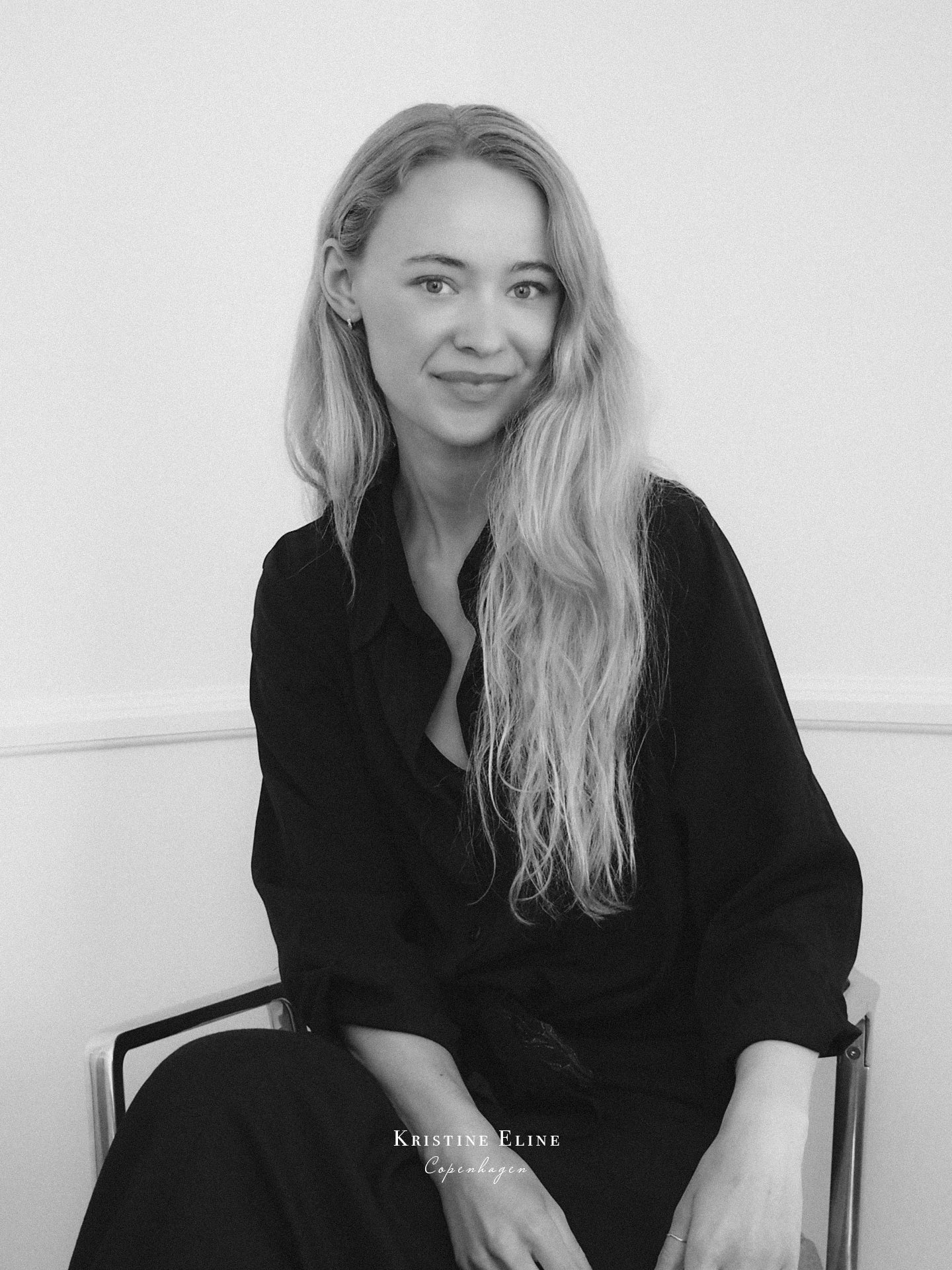Black and white portrait of the Co-Founder of Kristine Eline with long hair sitting on a chair, with logo on top.