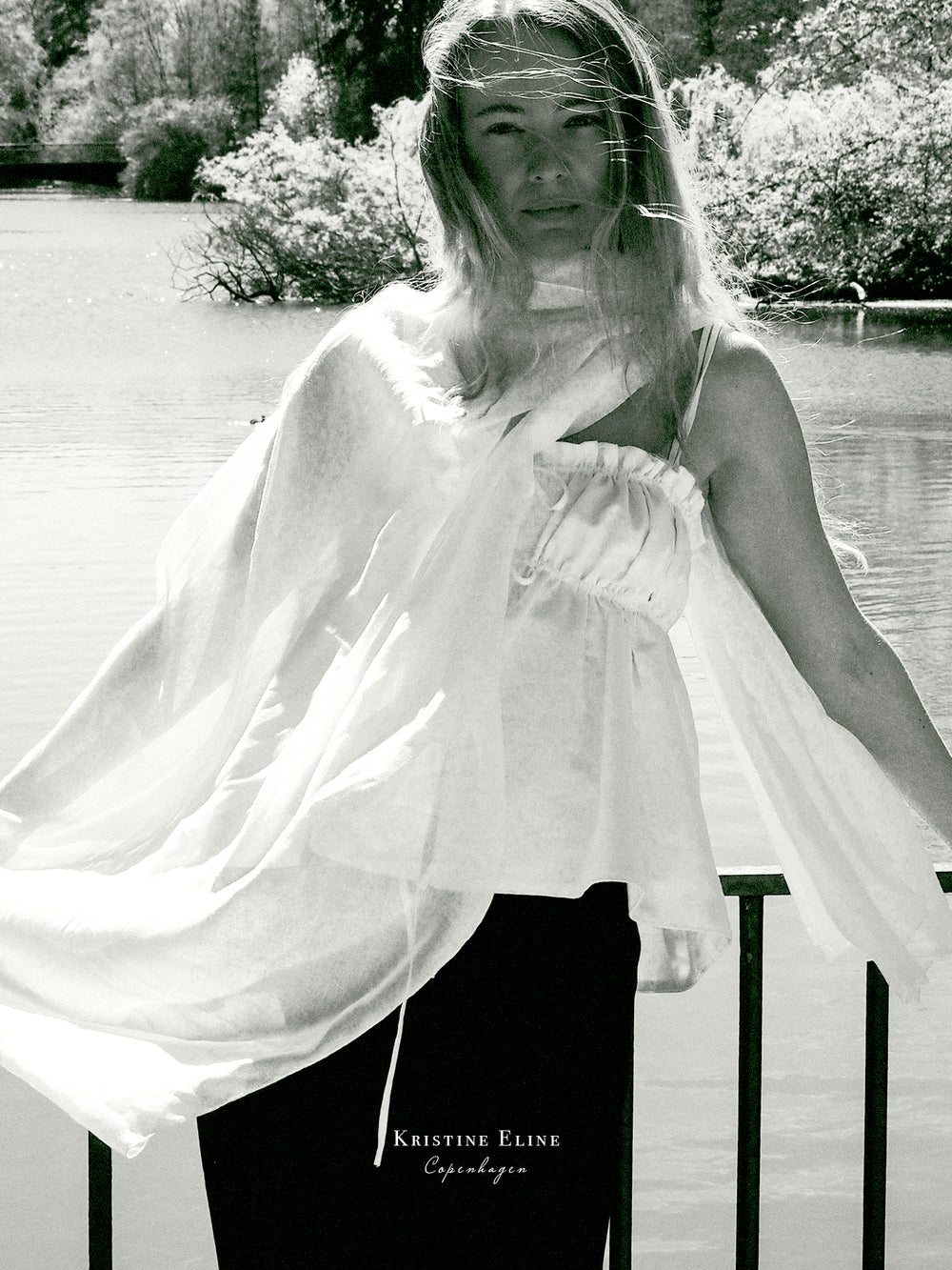 Woman in a flowing Ivory top standing outdoors by a river of water with 'Kristine Eline' logo.