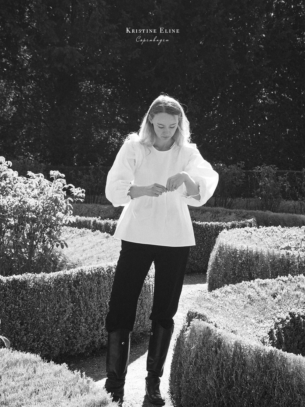 Girl standing in a garden with manicured hedges, wearing a white blouse. withpuffed sleeves, black pants and black high-knee boots.